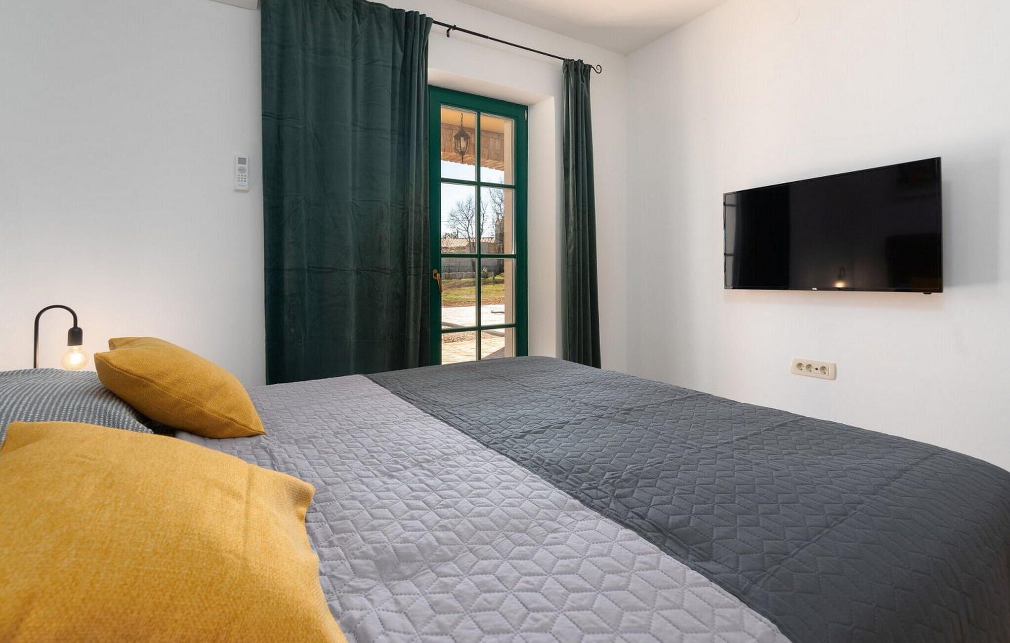 A bedroom with a bed, a TV, a window with curtains, and a bedside lamp.
