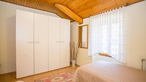 Une chambre avec un plafond en bois, une grande armoire, un lit recouvert d’une couvre-lit de couleur claire, une fenêtre ornée de rideaux légers et un vase décoratif contenant des branches.