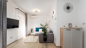 Een moderne woonkamer met een grijze bank, een klein wit tafeltje, een plant, een houten kast en een wandklok.