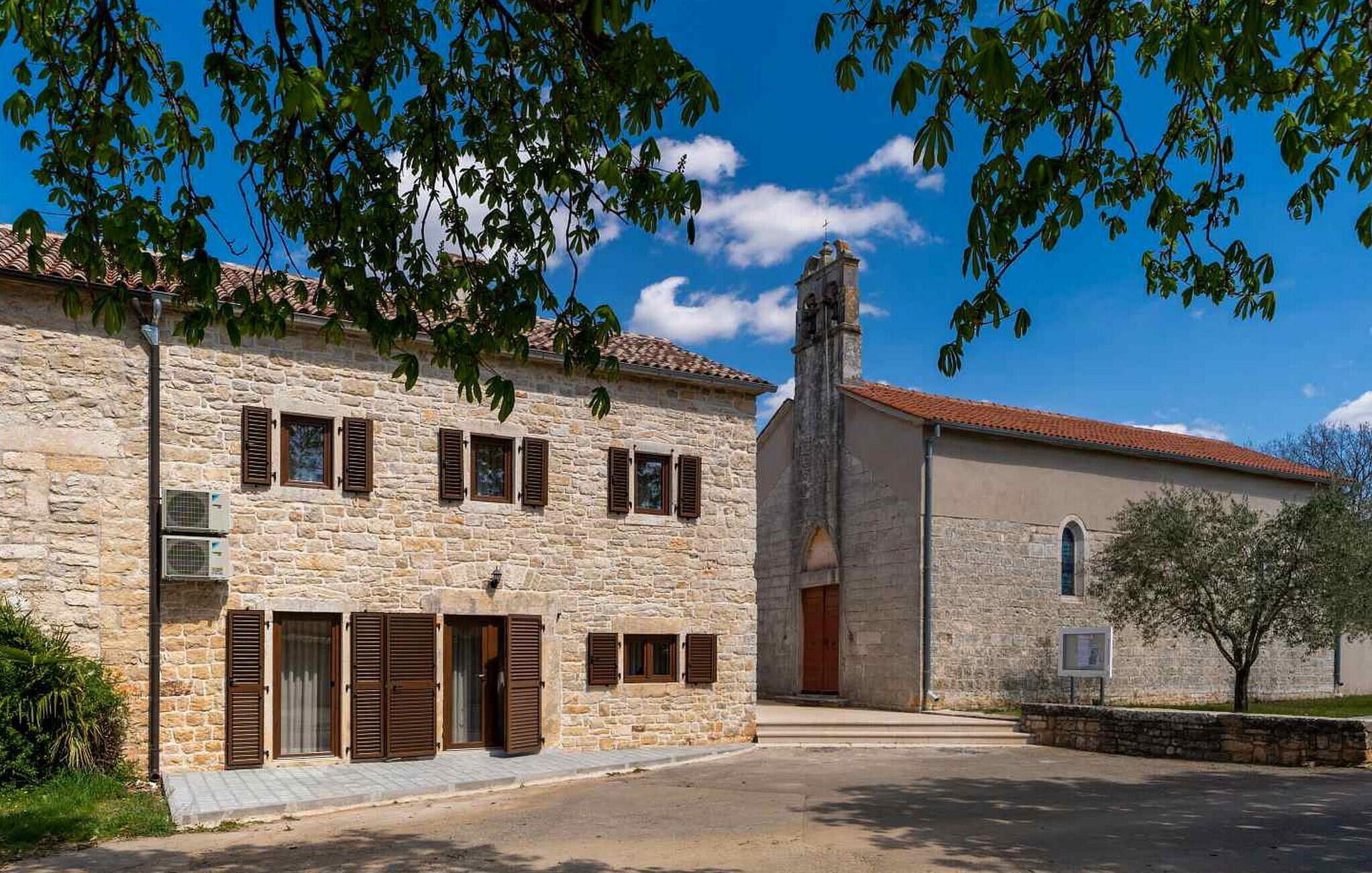 Um edifício de pedra com várias janelas e persianas, uma igreja com um campanário, e um céu azul claro.