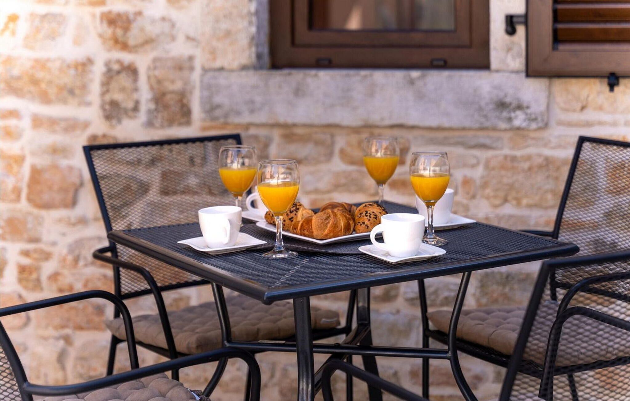 Une table mise pour le petit-déjeuner, avec des croissants, du jus d’orange et des tasses de café, entourée de chaises.