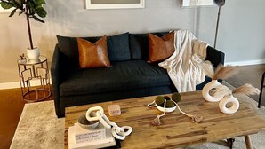 A living room with a dark sofa, two framed pictures, a potted plant, a coffee table with decorative items, and a lamp. - Located in the heart of Houston! Easy access to freeways, sports arenas, & more! (Houston)