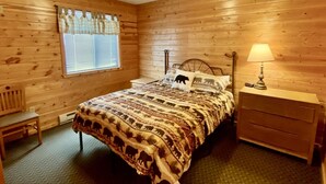 A wooden-paneled room with a bed, a nightstand, a lamp, a chair, and a window with blinds. - Cabin 9 Lakefront Family Resort (Hines)