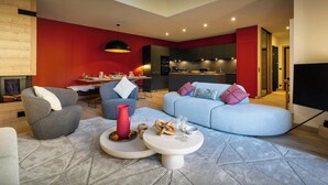 A modern living room with a sofa, armchairs, and a coffee table. There is a dining area with a table and chairs, and a kitchen in the background.