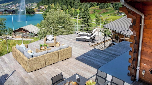 A wooden deck with outdoor furniture, a dining table with chairs, and a view of a lake and mountains in the background.