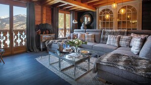 A cozy living room with a wooden ceiling, a sofa set, a glass coffee table, and a view of the mountains.