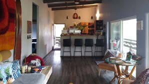 A spacious living area with a kitchen in the background, wooden flooring, and a large window providing natural light.