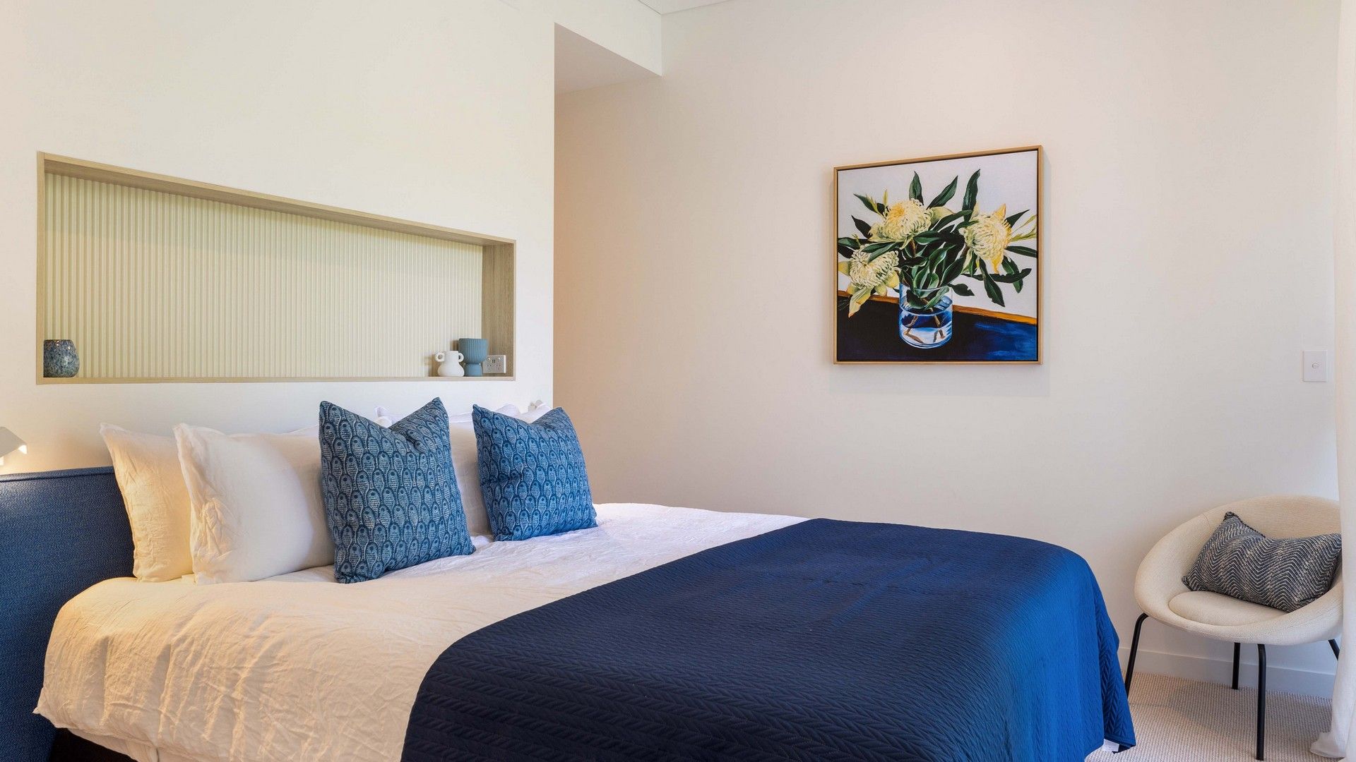 Une chambre à coucher comprenant un lit, des oreillers, une table de chevet, une chaise et une œuvre d’art accrochée au mur.