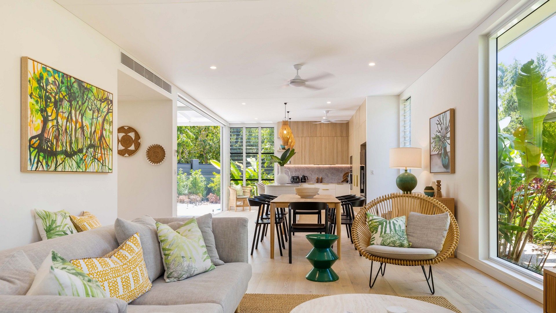 A modern living room with a sofa, dining table, chairs, and a rattan chair. There is a kitchen visible through an open doorway, and the room has large windows.