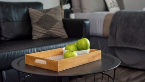 Een houten dienblad met groene appels op een ronde, zwarte tafel in een woonkamer met een donkere leren bank en een kussen met een patroon.