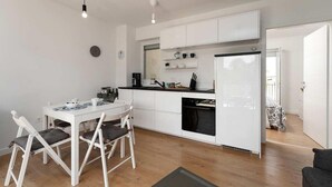 Une cuisine compacte avec des meubles blancs, un plan de travail noir et un réfrigérateur. On y trouve également un coin repas avec une table et des chaises blanches.