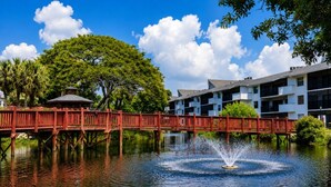 A red bridge over water with a fountain, surrounded by greenery and buildings. - Park Shore Resort-Top Floor BLDGC #221*Peaceful* (Park Shore Resort)