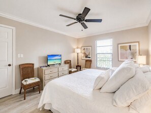 Une chambre à coucher comprenant un lit, un ventilateur de plafond, un téléviseur, une chaise, une commode et une fenêtre avec des stores.