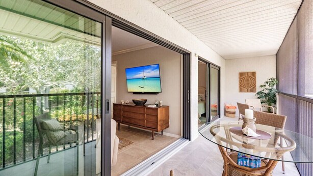 A modern living room with a glass dining table, wicker chairs, a wooden cabinet, and a flat-screen TV displaying a scenic view. - Park Shore Resort-Top Floor BLDGC #221*Peaceful* (Park Shore Resort)