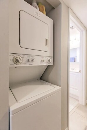 Un lavadero pequeño con lavadora y secadora de carga frontal, un estante sobre la secadora y una puerta que da a otra habitación.