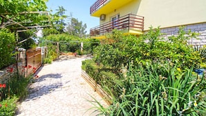 A pathway leading to a yellow building with a balcony, surrounded by lush greenery and a stone wall.