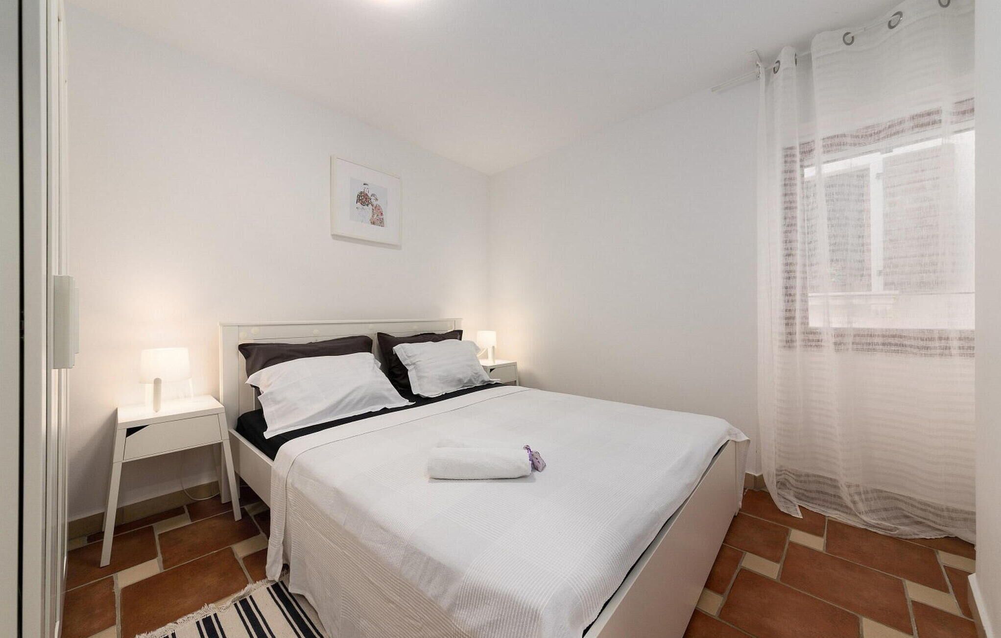 Un dormitorio ordenado con una cama, dos mesitas de noche, una lámpara de noche, un cuadro enmarcado en la pared y una ventana con cortinas traslúcidas.
