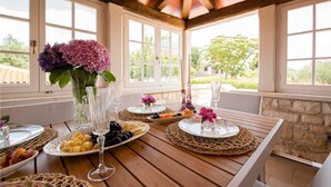 A covered outdoor dining area with a wooden table set for a meal, surrounded by large windows.
