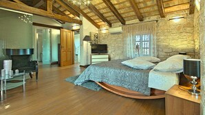 A spacious bedroom with a curved bed, wooden beams, stone walls, and a chandelier.