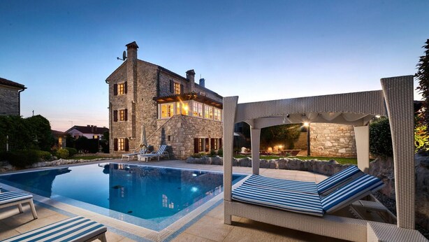 A stone house with a swimming pool, lounge chairs, and a pergola with lights.