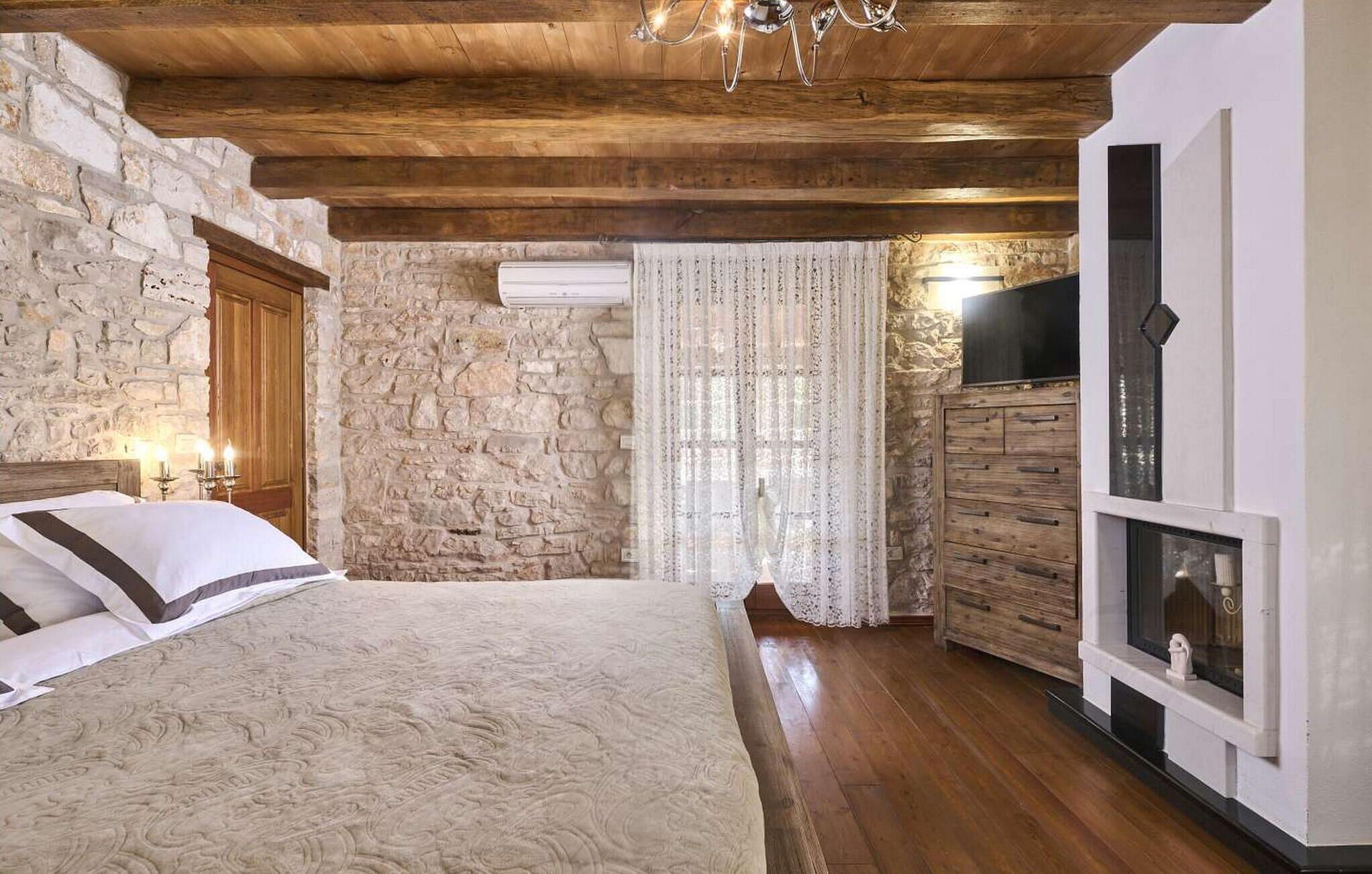 A bedroom with a stone wall, wooden ceiling, a bed with white and brown bedding, a wooden dresser, and a TV mounted on the wall.