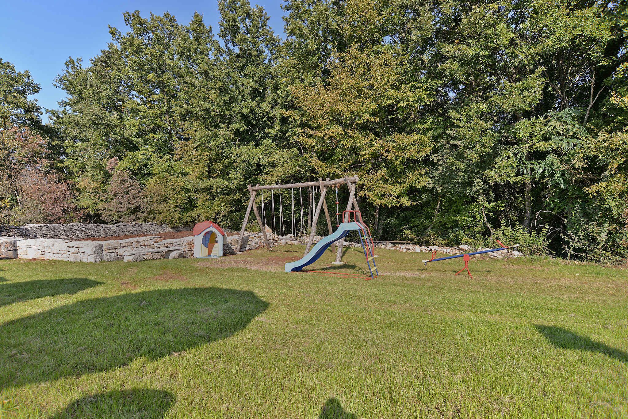 A playground with a swing set and slide, surrounded by trees and a stone wall.