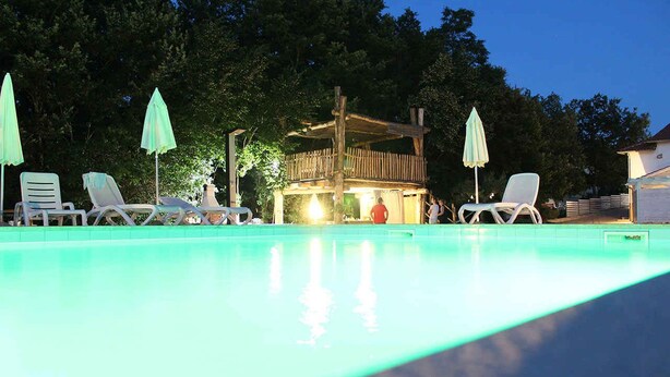 Piscina con sillas reclinables y sombrillas, rodeada de árboles y una terraza de madera.