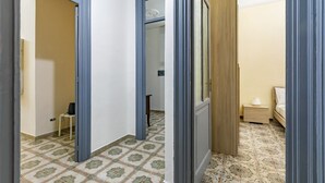 A hallway with patterned floor tiles, a door leading to a bedroom with a bed and nightstand, and a chair in the background.