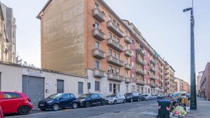 Un immeuble résidentiel à plusieurs étages, doté de balcons, avec des voitures garées et une poubelle sur le trottoir.