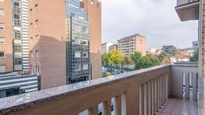 Un balcon donnant sur des immeubles modernes, un sol carrelé et une rambarde en métal.