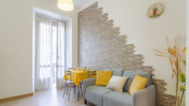Un salon avec un canapé gris, des coussins jaunes, une table à manger avec des chaises et un mur en pierre.