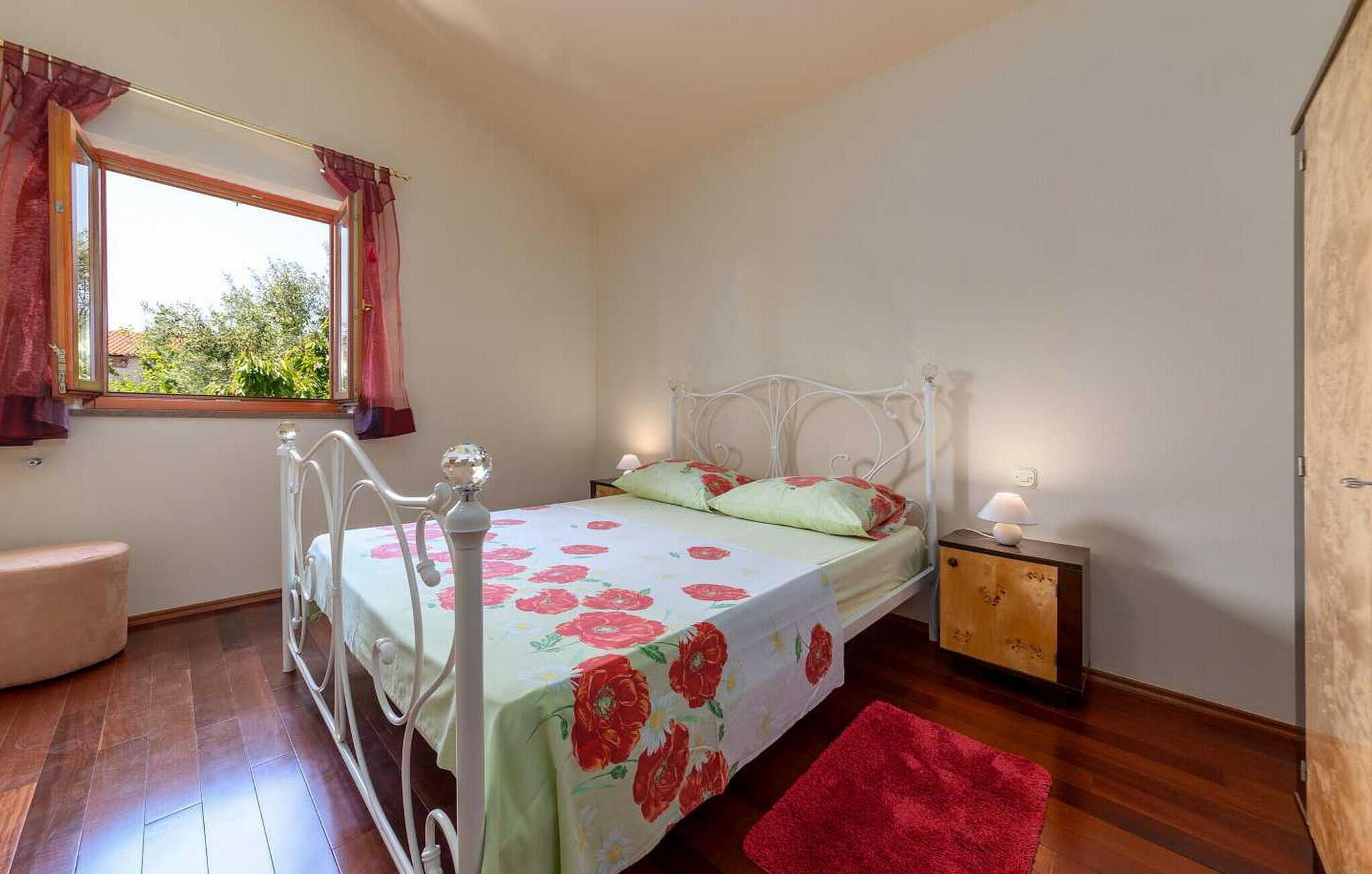 A bedroom with a bed, a nightstand, a window with curtains, and a red rug.