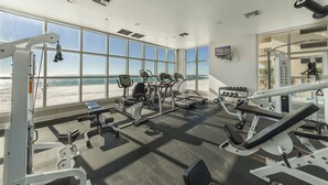 A gym with various exercise equipment, large windows with a beach view, and a television mounted on the wall.