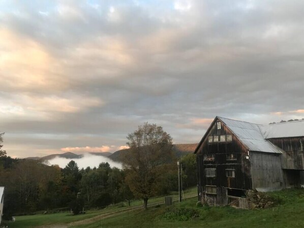 A rustic barn with a sloped roof and a large window, surrounded by a lush green landscape and a cloudy sky.