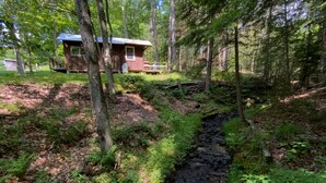 A rustic cabin surrounded by dense forest, a small stream, and a wooden deck. - The Creek Cabin at Sky Hollow, The Creek Cabin at Sky Hollow (Rochester)