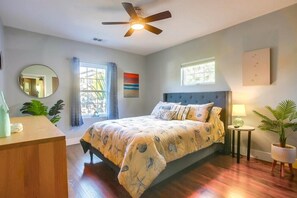 A bedroom with a bed, a ceiling fan, a window, a bedside table, and a potted plant.