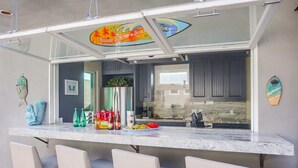 A kitchen area with a marble countertop, bar stools, a refrigerator, and a surfboard decoration. - Amazing Beach House! 2 Yards w/ Hot Tub + Fire Pit (San Diego)