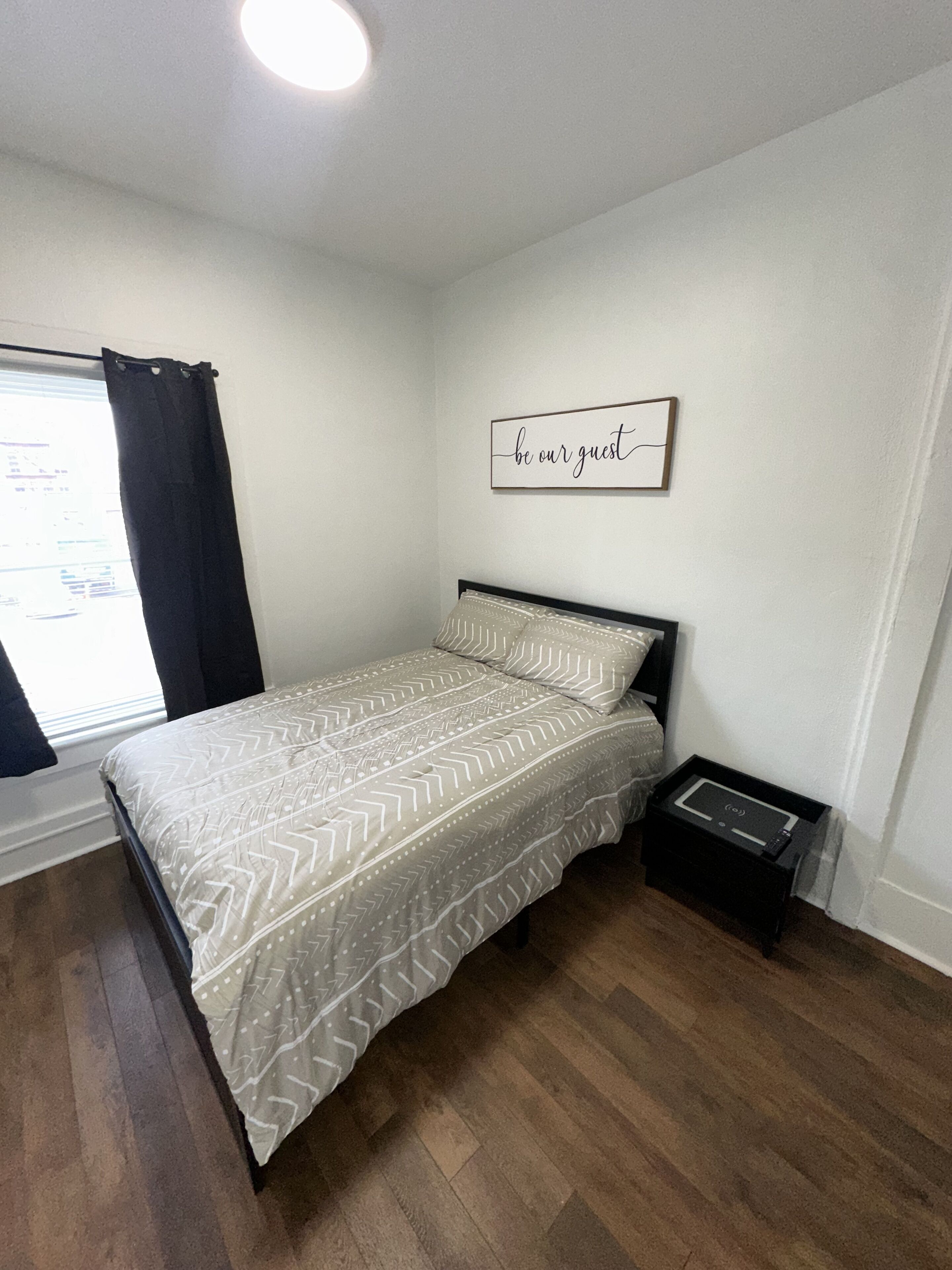 A bedroom with a bed, a bedside table, a wall-mounted artwork, a window with curtains, and a ceiling light.