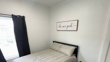 A bedroom with a bed, a bedside table, a wall-mounted artwork, a window with curtains, and a ceiling light.