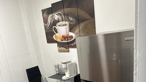 Ett kompakt kök med ett kylskåp i rostfritt stål, ett matbord med glasskiva och två svarta stolar samt ett väggmonterat konstverk med kaffetema.
