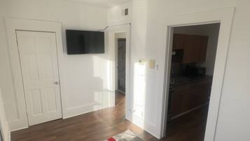 Amplia sala de estar con pisos de madera, un sofá negro, un tapete rojo y gris, un ventilador de techo y un televisor de pantalla plana montado en la pared.