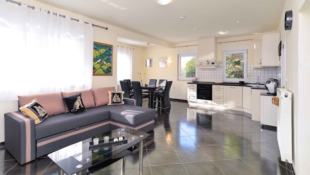 Una sala de estar moderna con un sofá modular, una mesa de centro de vidrio y un comedor con mesa y sillas negras. La cocina cuenta con gabinetes blancos y una encimera negra.