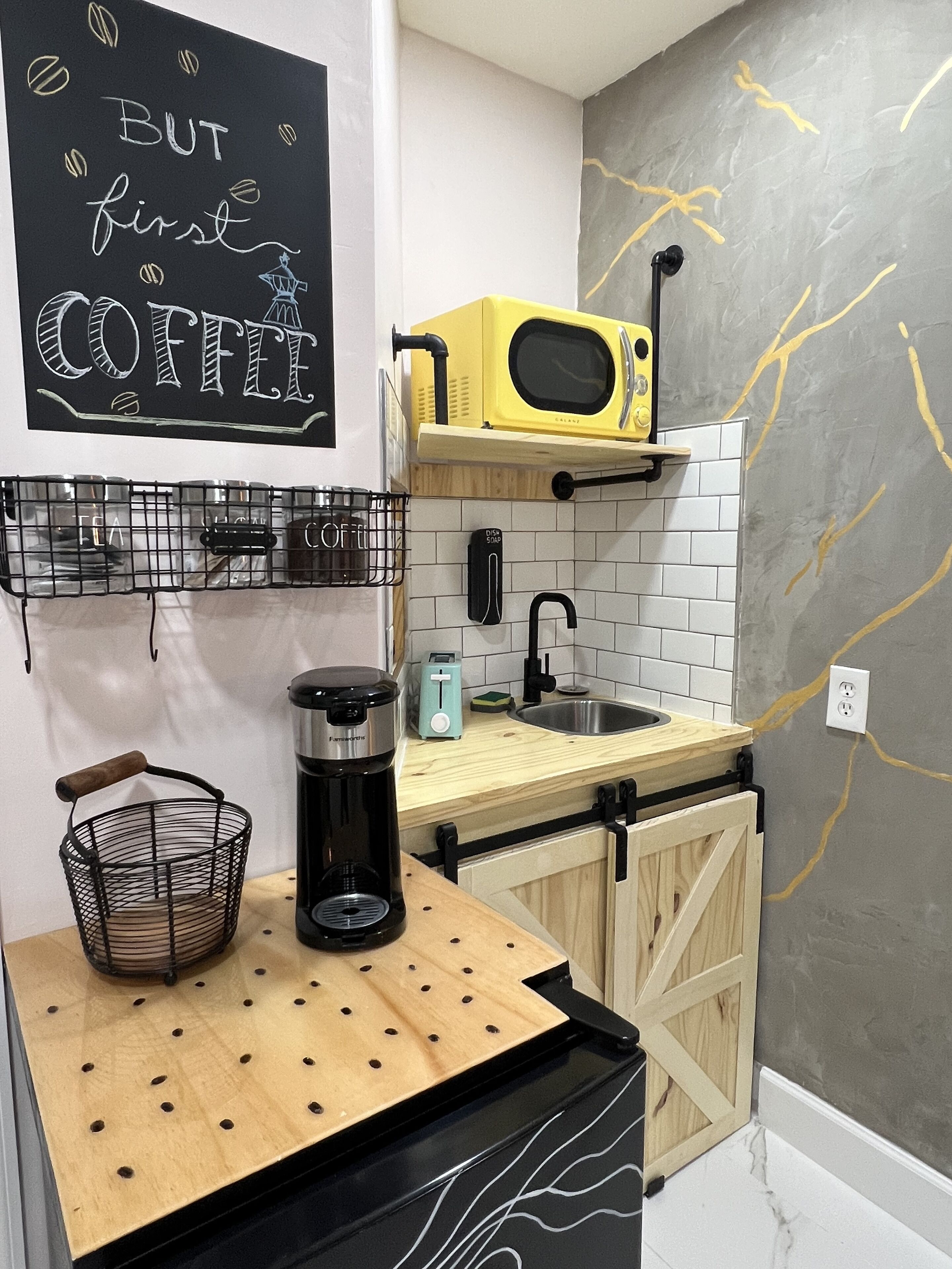 Cocina compacta con microondas, cafetera y una pizarra con la inscripción 'BUT first COFFEE'.