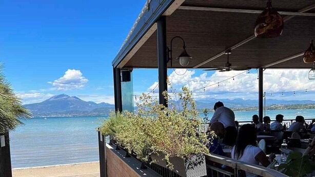 A seaside restaurant with outdoor seating overlooking a calm sea and distant mountains.