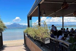 Un restaurante frente al mar con terraza al aire libre, con vistas a un mar tranquilo y montañas lejanas.