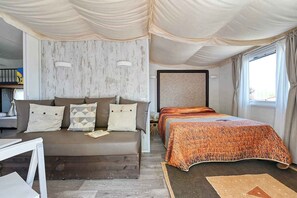 A bedroom with a sofa bed, a separate bed with an orange patterned bedspread, a wooden headboard, and a large window with curtains. - Mobilehome with air conditioning, terrace and pool (Peschiera del Garda)