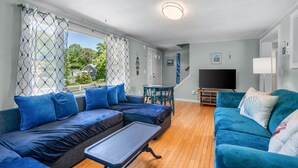 A living room with a blue sofa, a blue armchair, and a TV. There is a window with curtains, a door, and a sign that says "Welcome to Our Home".