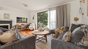 A living room with wicker furniture, a fireplace, a large window with curtains, and a view of greenery outside.