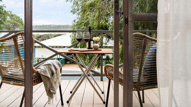A balcony with wicker chairs, a table with wine glasses and a bottle, and a view of a bridge and greenery.