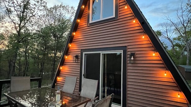 A triangular wooden cabin with a glass table and chairs on a deck, surrounded by trees. - Secluded A frame cabin close Dells, BRAND NEW HOT TUB, firepit, grill. (Oxford)